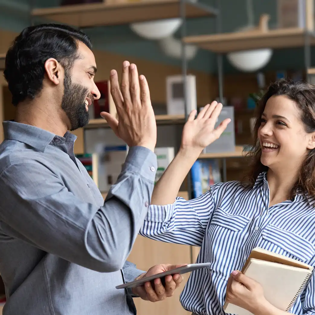 A man and women are about to high-five. The man is holding a tablet, and the woman is holding a notepad with their other hands.