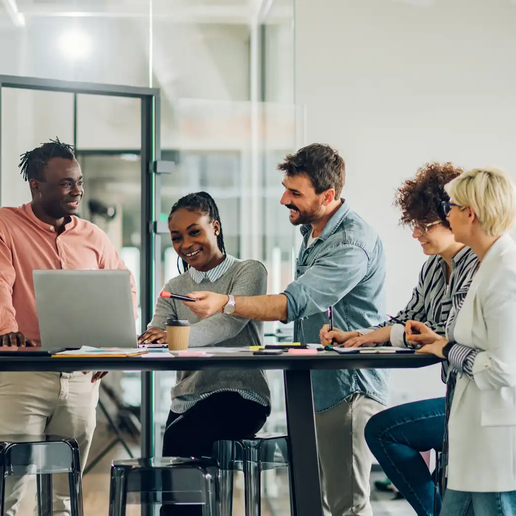 Five individuals crowded around a table discussing what they see on their laptops while others are writing down notes and contributing to the conversation.