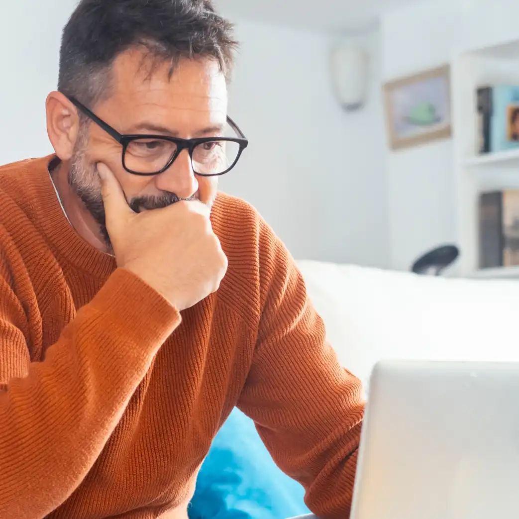 A man wearing glasses and an orange sweater appears to be thinking and looking at a laptop