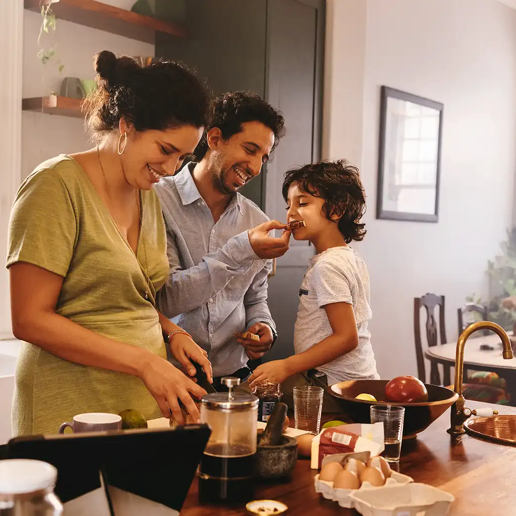 A mom, dad, and son eating a snack in their kitchen. The father is feeding the kid while the mother is cutting a snack into smaller pieces. They are all smiling.