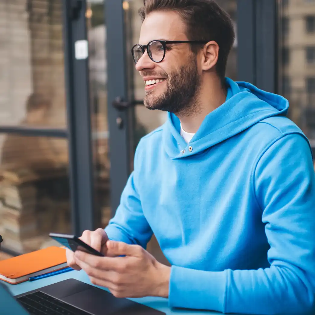 A man wearing a blue hoodie and glasses smiling with his phone in his hands.