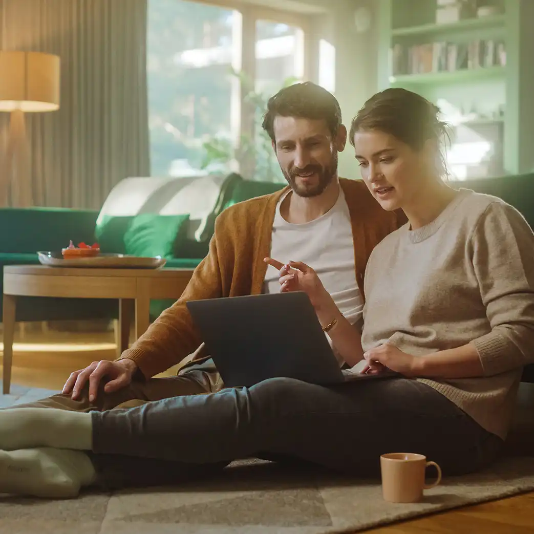 A man and woman sitting on the floor discussing what they see on a laptop screen.