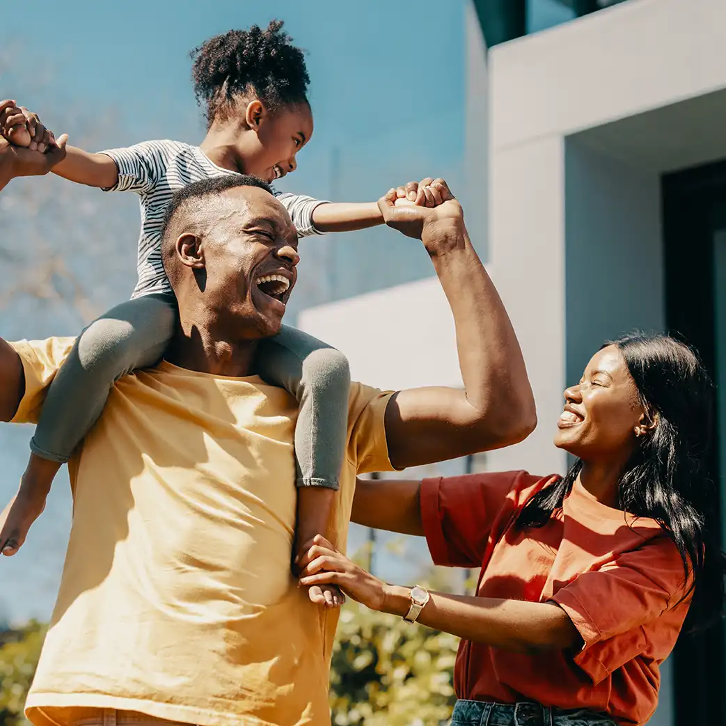 A woman, man, and daughter are pictured in a group photo. The daughter is on the man's shoulders and the women is holding on to her daughters foot and man's back. Everyone is smiling and laughing.