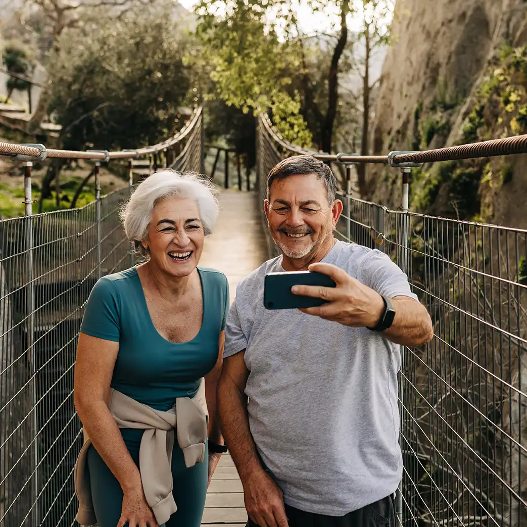 A older man and woman smiling and taking a picture on a walking bridge during their hike.