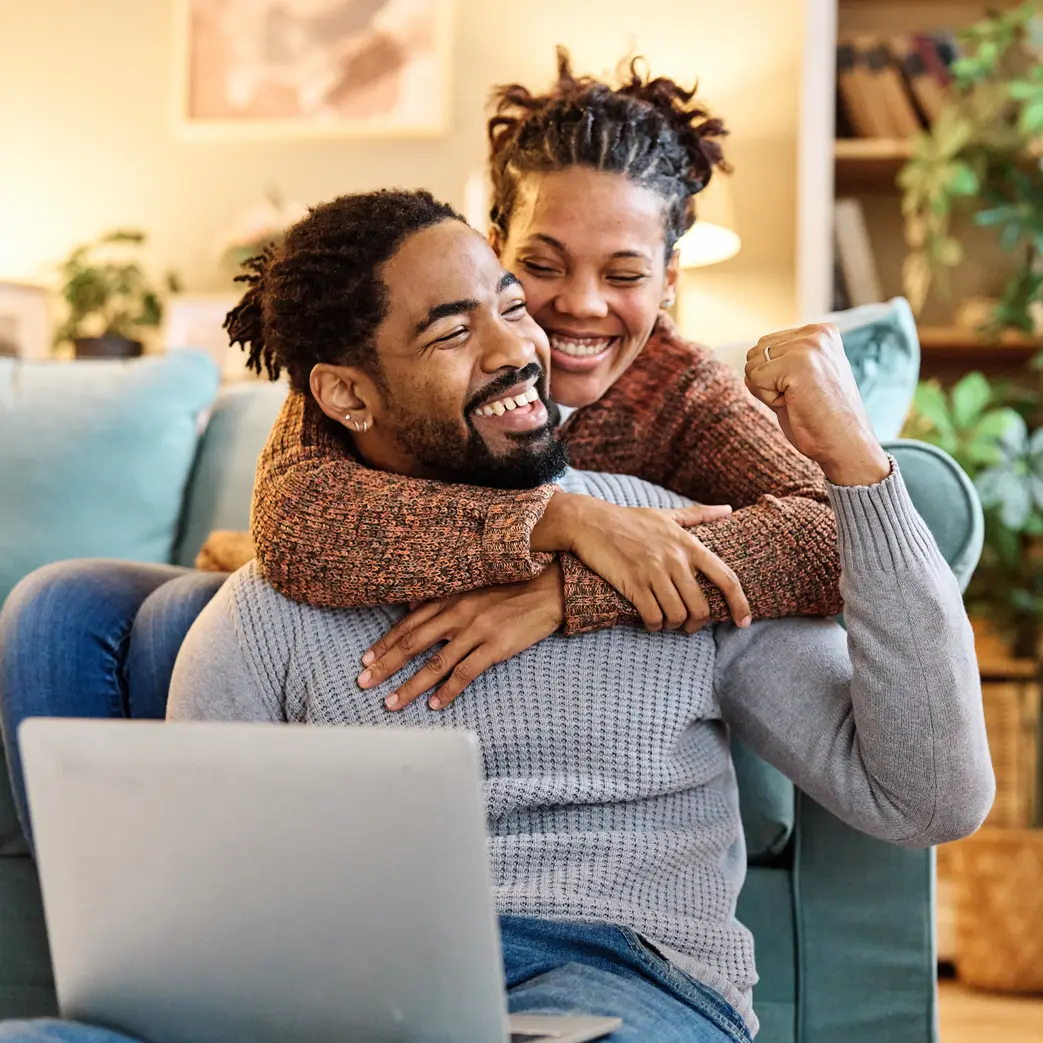 A women hugging her man from behind around his shoulders while she sits on the couch, and he is sitting on the floor. They are both smiling. The man is holding a laptop and fist bumping the air.